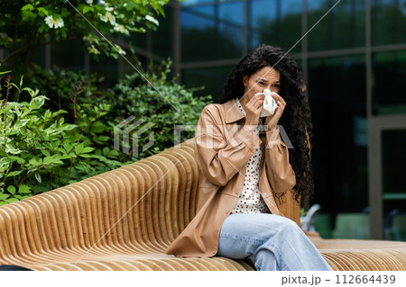 A woman sits on an outdoor bench, blowing her nose with a tissue, possibly suffering from cold or allergies, in a natural, urban setting. 112664439