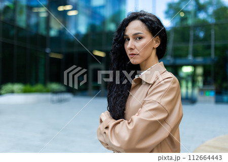 A professional woman in a beige shirt crosses her arms confidently outside modern office buildings. A professional woman in a beige shirt crosses her arms confidently outside modern office buildings. 112664443