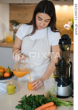 Woman pouring orange homemade juice into glass. Female in apron in the kitchen. Fruits and vegetables on the table. Juicer. Squeezer Woman pouring orange homemade juice into glass. Female in apron in the kitchen. Fruits and vegetables on the table. Juicer. Squeezer 112665288