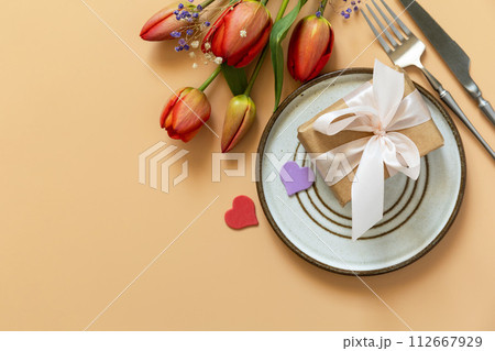 Beautiful table setting with gift box, decorative hearts and tulips flowers on pastel peach background. Beautiful table setting with gift box, decorative hearts and tulips flowers on pastel peach background. 112667929