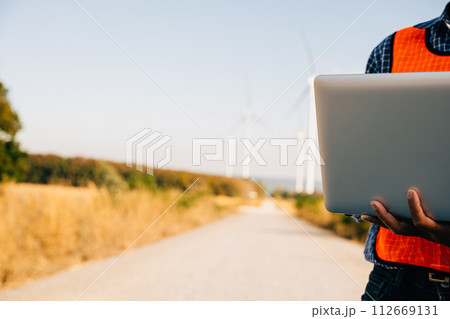 Engineer laptop in hand assesses wind turbines. Service technician ensures efficiency. Focused on innovation quality and landscape development in windmill service. 112669131