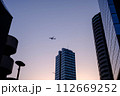夕暮れにビルの間を飛ぶ飛行機 112669252