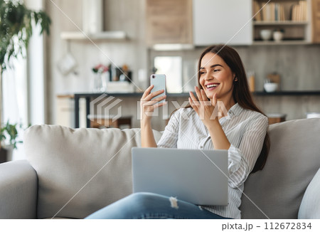 AI-generated content. A woman is sitting comfortably on a couch, engaging in a phone conversation while simultaneously using a laptop. Her focus is on the electronic devices in her hands 112672834