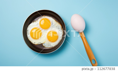 Two fried eggs in a pan, wooden spoon centerpiece, set against a blue background, Ai Generated. 112677838