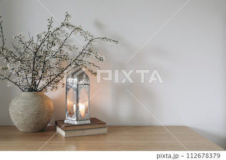 White flowers, blooming prunus tree branches in vase. Glowing Moroccan lantern on books. Wooden table. White wall background. Iftar dinner. Ramadan Kareem greeting card. Eid ul Fitr muslim holiday. White flowers, blooming prunus tree branches in vase. Glowing Moroccan lantern on books. Wooden table. White wall background. Iftar dinner. Ramadan Kareem greeting card. Eid ul Fitr muslim holiday. 112678379
