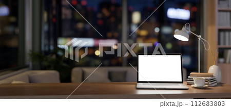 A laptop mockup on a wooden desk in a contemporary home office at night illuminated by dim lights. 112683803