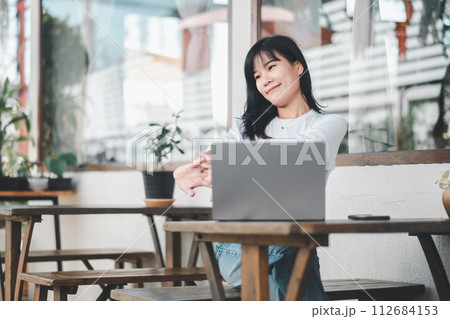 Relaxed young woman stretching while working on a laptop in a casual outdoor cafe setting, enjoying a work-life balance. 112684153