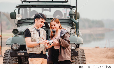 Young couple stands together outdoors, sharing a moment over a vintage camera with a car in the background. Young couple stands together outdoors, sharing a moment over a vintage camera with a car in the background. 112684190