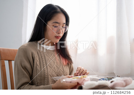 A young Asian woman is focusing on threading a pattern on an embroidery frame, hand sewing on cloth. 112684256