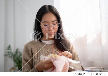 A young Asian woman is focusing on threading a pattern on an embroidery frame, hand sewing on cloth. 112684258