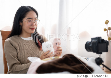 A cheerful young Asian female content creator showing her cute handmade plushie to the camera. A cheerful young Asian female content creator showing her cute handmade plushie to the camera. 112684318