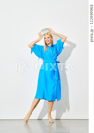 Cheerful woman in a blue shirtdress holds a silver purse over her head 112690968