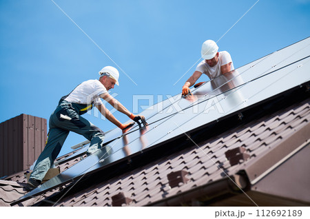 Workers building solar panel system on rooftop of house. Two men installers in helmets installing photovoltaic solar module outdoors. Alternative, green and renewable energy generation concept. Workers building solar panel system on rooftop of house. Two men installers in helmets installing photovoltaic solar module outdoors. Alternative, green and renewable energy generation concept. 112692189