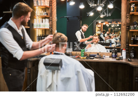 Regular client enjoying his drink while getting haircut. Professional hairdresser making stylish haircut, Luxury barbershop offering drink. 112692200