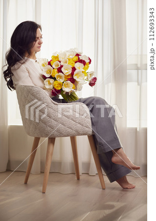Beautiful woman with spring flowers tulips in hands sitting on chair near window. Women's Day Beautiful woman with spring flowers tulips in hands sitting on chair near window. Women's Day 112693343
