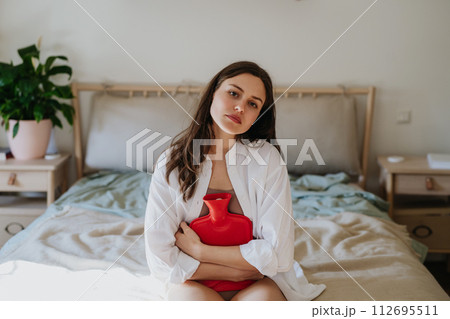 Woman at home suffering from menstrual pain, having cramps. Woman warming lower abdomen with a hot water bottle, endometriosis, and conditions causing pain in tummy. 112695511