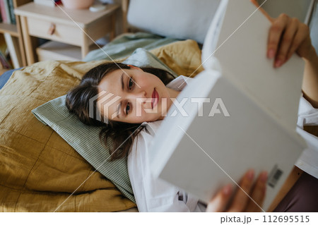 Beautiful woman reading book in bed, cozy, warm and contented feeling. Freedom in story, weekend activity for single woman. 112695515