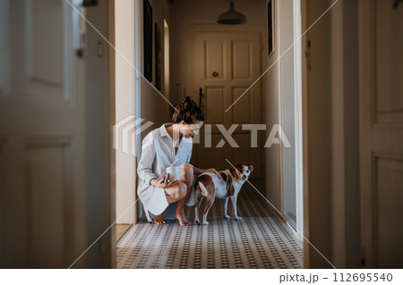 Woman in pajamas playing with her cute dog, drinking cup of morning coffee. Weekend morning relaxation at home. Hygge lifestyle. Woman in pajamas playing with her cute dog, drinking cup of morning coffee. Weekend morning relaxation at home. Hygge lifestyle. 112695540
