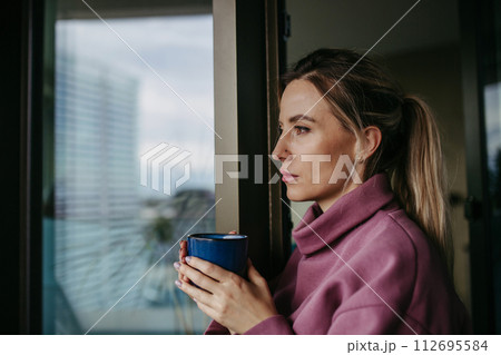 Woman enjoying cup of hot tea, coffee at home after work, standing by window wearing pink hoodie. Work-life balance for healthcare worker. 112695584