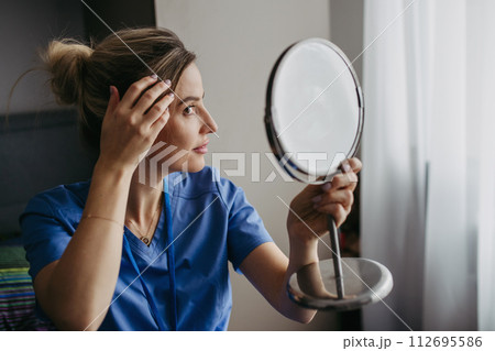Female nurse or doctor getting ready for work in the morning, brushing hair, making hairstyle in bedroom. Work-life balance for healthcare worker. 112695586