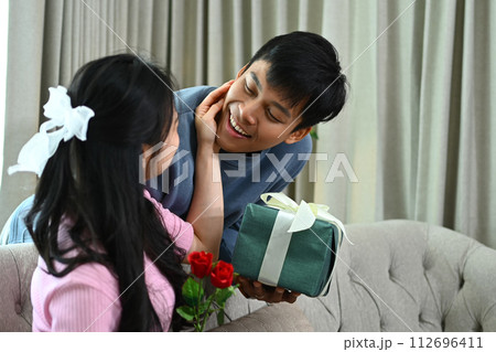 Smiling man surprising his girlfriend with a gift box on couch at home. Love, family and celebration concept 112696411