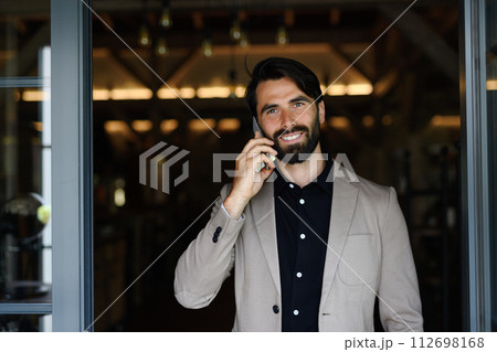 Portrait of mature businessman in suit jacket standing indoors in modern office, making phone call. 112698168