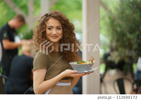 Portrait of a beautiful waitress holding plate with food, bowl with salad. Server standing on restaurant terrace in an apron. 112698172