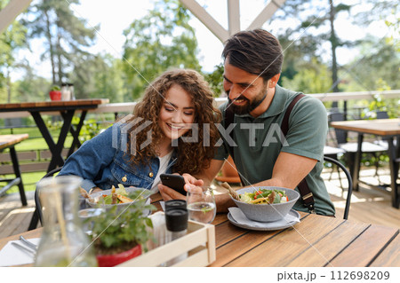 Old friends sitting on restaurant terrace, meeting after long time, looking at old photos in smatphone. Friends enjoying springtime, having lunch outdoors. 112698209