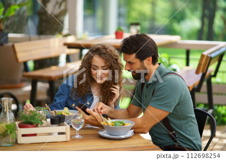 Old friends sitting on restaurant terrace, meeting after long time, looking at old photos in smatphone. Friends enjoying springtime, having lunch outdoors. Old friends sitting on restaurant terrace, meeting after long time, looking at old photos in smatphone. Friends enjoying springtime, having lunch outdoors. 112698254