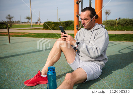 Young athletic man sitting on the sports ground and checking health sport mobile app on smartphone after outdoor workout 112698526