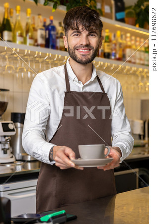 Barista standing by the counter in coffee shop holding cup of coffee for client Barista standing by the counter in coffee shop holding cup of coffee for client 112698622
