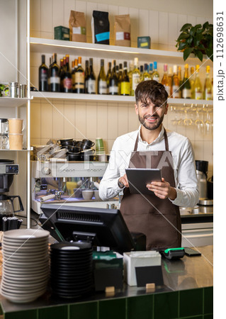 Bearded man bartender standing with digital tablet at coffee shop counter Bearded man bartender standing with digital tablet at coffee shop counter 112698631