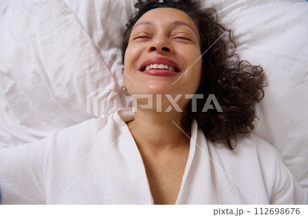 Close-up portrait of a smiling curly young woman smiling while waking up in the morning , lying on the bed Close-up portrait of a smiling curly young woman smiling while waking up in the morning , lying on the bed 112698676