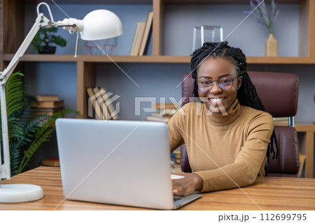 Woman remote worker in glasses with dreadlocks sitting at workplace with laptop in home office 112699795