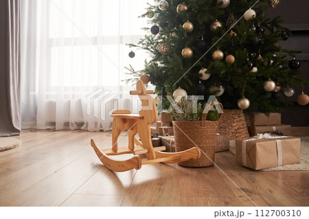 View of wooden rocking rabbit in the center of living room with decorated christmas tree, wicker basket with spruce branches, gifts in craft paper with copy space. View of wooden rocking rabbit in the center of living room with decorated christmas tree, wicker basket with spruce branches, gifts in craft paper with copy space. 112700310
