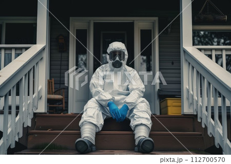 A disinfection service worker in a protective suit and gas mask sits on the porch of the house 112700570