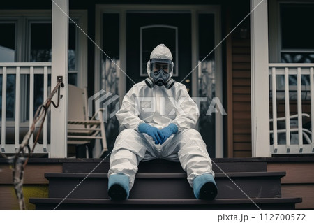 A disinfection service worker in a protective suit and gas mask sits on the porch of the house 112700572