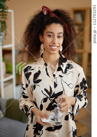 Vertical portrait of young Black woman with curly hair holding tiny watering can and smiling at camera Vertical portrait of young Black woman with curly hair holding tiny watering can and smiling at camera 112703291