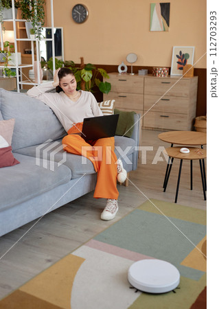 Vertical full length portrait of young woman using laptop on couch and relaxing with robot vacuum cleaner mopping floors Vertical full length portrait of young woman using laptop on couch and relaxing with robot vacuum cleaner mopping floors 112703293