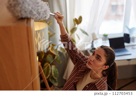 High angle portrait of young woman dusting tall shelves and hard to reach places while doing Spring cleaning at home High angle portrait of young woman dusting tall shelves and hard to reach places while doing Spring cleaning at home 112703453