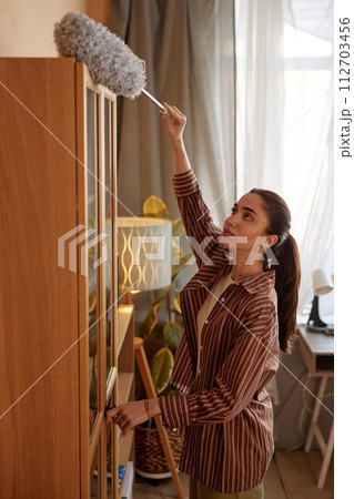 Vertical portrait of young woman dusting top of tall cupboard while doing Spring cleaning at home 112703456