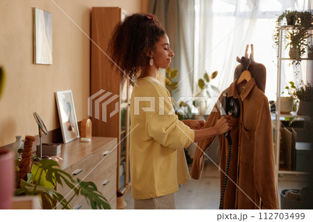 Side view portrait of young woman with curly hair steaming velvet jacket while taking care of clothes in cozy home 112703499