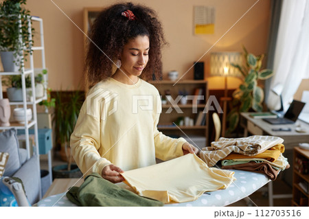 Waist up portrait of smiling young woman with big curly hair folding clothes on ironing board at home copy space 112703516