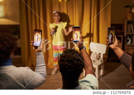 Black little girl performing in home concert or talent show with parents holding smartphone and recording video copy space 112704878