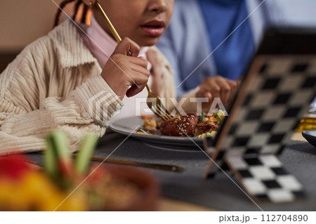Close up of little Black girl eating dinner at table and watching videos online copy space 112704890