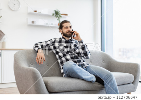 Young man in casual clothes talking on smartphone while sitting on sofa. 112706647