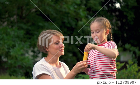 Little girl and mother blow soap bubbles together in green city park. Mother with joyful little girl plays soap bubbles walking in park. Little girl and mother rest blowing soap bubbles in garden Little girl and mother blow soap bubbles together in green city park. Mother with joyful little girl plays soap bubbles walking in park. Little girl and mother rest blowing soap bubbles in garden 112709907