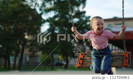 Laughing cute baby girl swinging on swing at public city summer park enjoy outdoor leisure activity closeup. Smiling excitement female kid relaxing outside flying playground freedom positive emotion Laughing cute baby girl swinging on swing at public city summer park enjoy outdoor leisure activity closeup. Smiling excitement female kid relaxing outside flying playground freedom positive emotion 112709961