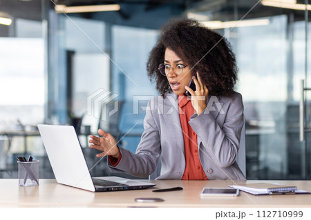 Terrified female talking by mobile phone while looking at portable computer screen with rounded eyes. Furious black woman getting desperate while receiving bad urgent news by business partner. Terrified female talking by mobile phone while looking at portable computer screen with rounded eyes. Furious black woman getting desperate while receiving bad urgent news by business partner. 112710999