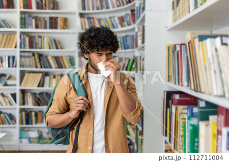 Unhealthy man in casualwear using paper napkin while sneezing and standing by aisle with collection of novels. Sick islamic male struggling with dust allergy in book shop while holding shoulder bag. 112711004